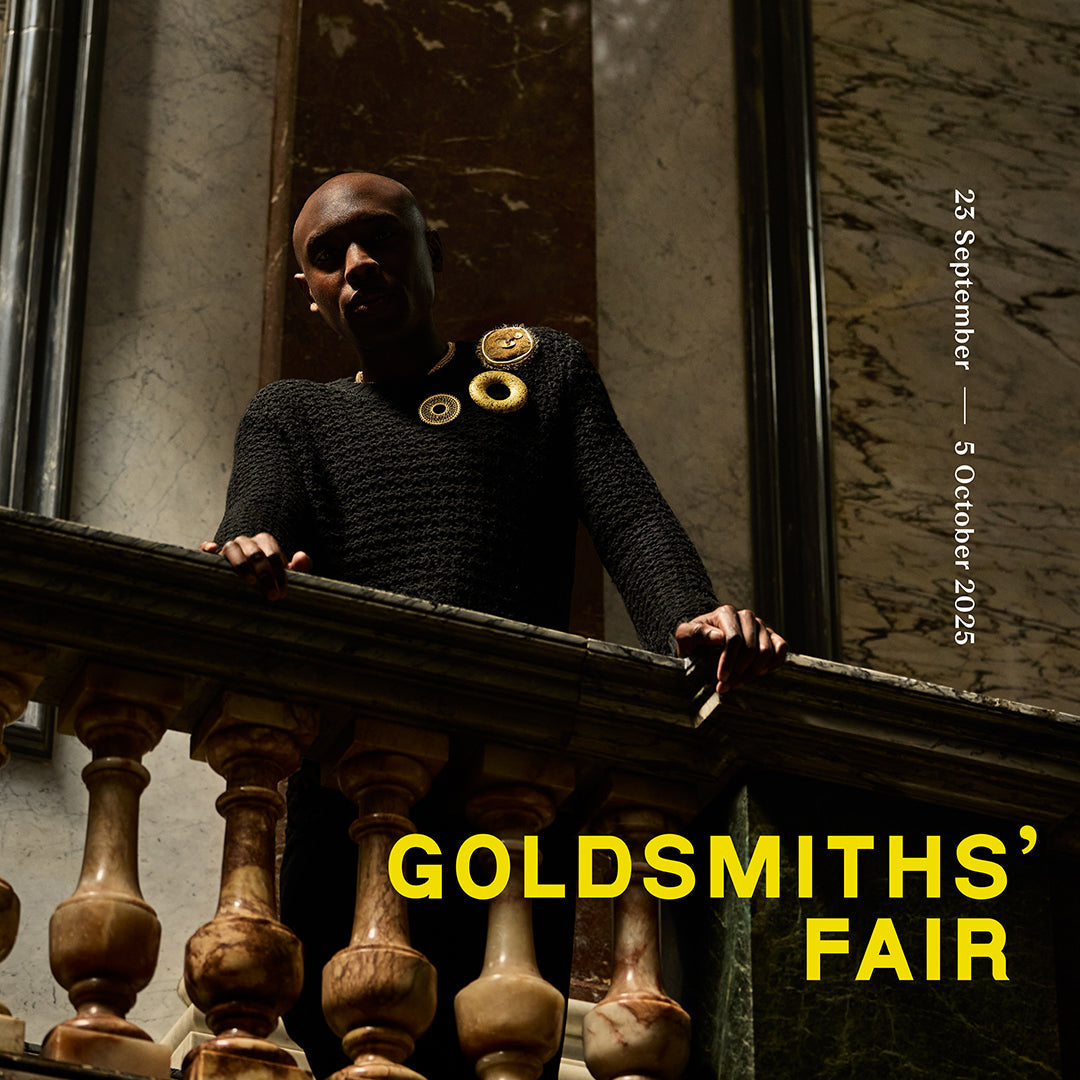 A person is standing on a balcony looking down, wearing three golden brooches, and the words Goldsmiths' Fair is written across the bottom